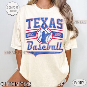 Pode incluir: Uma camiseta de cor creme com um gr&aacute;fico de beisebol vintage. O gr&aacute;fico apresenta um jogador de beisebol em um c&iacute;rculo azul com as palavras "Texas Baseball" em letras azuis e vermelhas. O texto "Est. 1835" tamb&eacute;m est&aacute; inclu&iacute;do em letras vermelhas.