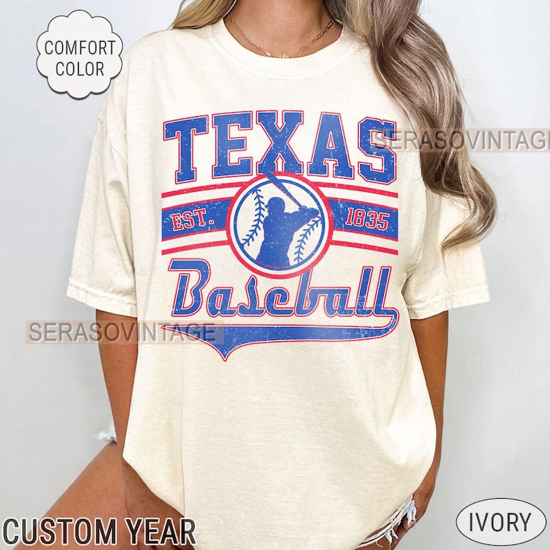Pode incluir: Uma camiseta de cor creme com um gr&aacute;fico de beisebol vintage. O gr&aacute;fico apresenta um jogador de beisebol em um c&iacute;rculo azul com as palavras "Texas Baseball" em letras azuis e vermelhas. O texto "Est. 1835" tamb&eacute;m est&aacute; inclu&iacute;do em letras vermelhas.