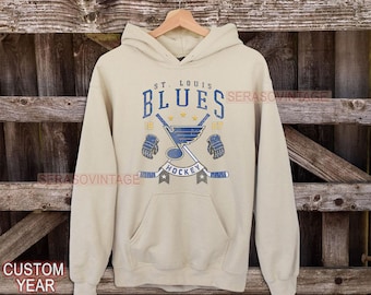 May include: A beige hoodie with a vintage St. Louis Blues hockey logo. The logo features a blue note with a hockey stick and a glove on each side. The text "St. Louis Blues Hockey" is printed below the logo.