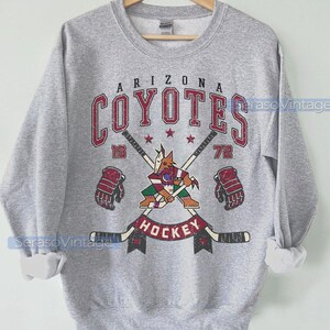 Puede incluir: Una sudadera gris con un gr&aacute;fico de estilo vintage del equipo de hockey de los Arizona Coyotes. El gr&aacute;fico presenta un coyote de dibujos animados sosteniendo un palo de hockey, con el nombre y el logotipo del equipo encima. El texto "19" y "72" est&aacute; a cada lado del gr&aacute;fico, y la palabra "Hockey" est&aacute; debajo.