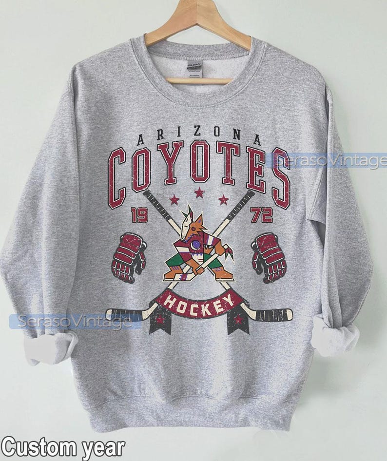 Puede incluir: Una sudadera gris con un gr&aacute;fico de estilo vintage del equipo de hockey de los Arizona Coyotes. El gr&aacute;fico presenta un coyote de dibujos animados sosteniendo un palo de hockey, con el nombre y el logotipo del equipo encima. El texto "19" y "72" est&aacute; a cada lado del gr&aacute;fico, y la palabra "Hockey" est&aacute; debajo.
