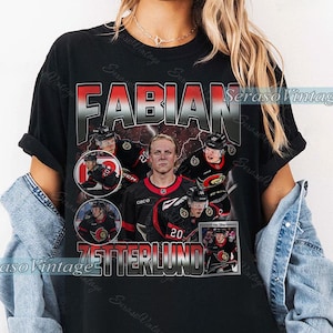 Puede incluir: Camiseta negra con un diseño de jugador de hockey. La camiseta presenta el nombre "FABIAN" en rojo y plata, y el nombre "ZETTERLUND". También se incluyen imágenes del jugador.