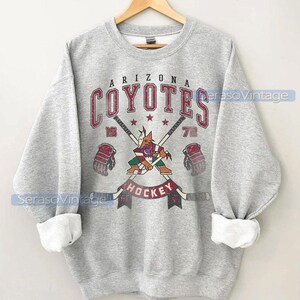 Puede incluir: Sudadera gris con un logotipo vintage de hockey de los Arizona Coyotes. El logotipo presenta un coyote mascota sosteniendo un palo de hockey con el nombre del equipo y los a&ntilde;os 19 y 72.