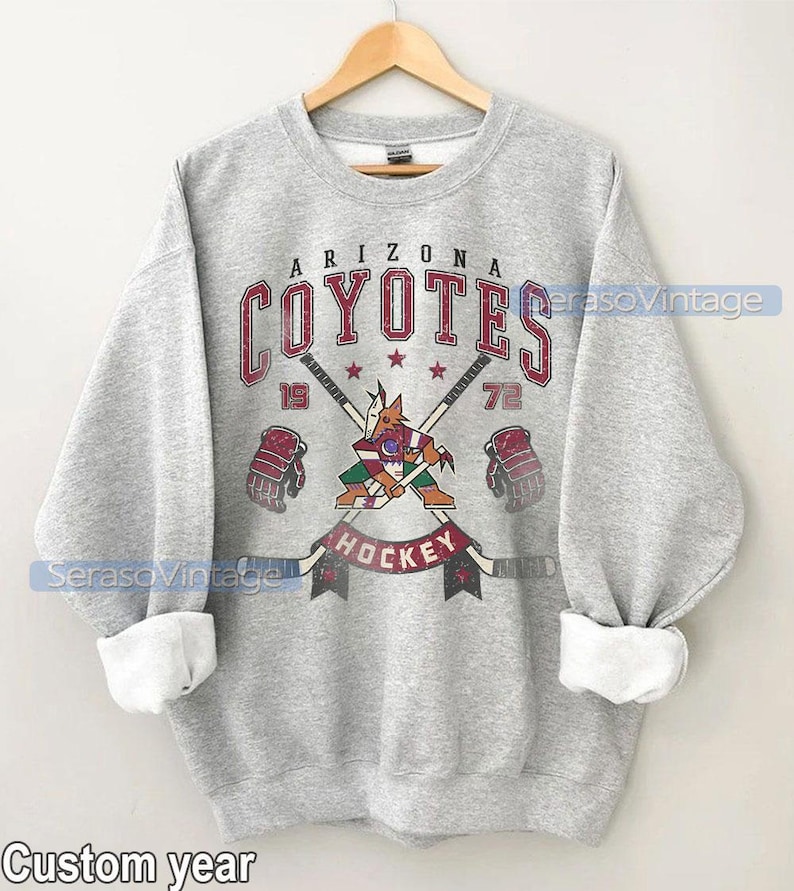 Puede incluir: Sudadera gris con un logotipo vintage de hockey de los Arizona Coyotes. El logotipo presenta un coyote mascota sosteniendo un palo de hockey con el nombre del equipo y los a&ntilde;os 19 y 72.