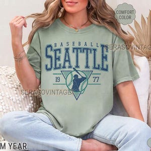 May include: A green t-shirt with a vintage baseball graphic. The graphic features a baseball player in a triangle with the text "BASEBALL SEATTLE 19 77".
