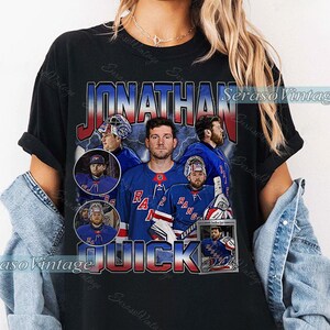 Può includere: T-shirt nero con grafica in stile vintage del giocatore di hockey Jonathan Quick con la maglia dei New York Rangers. Il design include il nome del giocatore e immagini di lui in azione. La maglia ha un aspetto invecchiato.