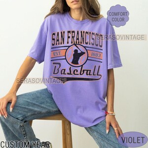 May include: A purple t-shirt with a vintage baseball graphic. The graphic features a baseball player in silhouette, a baseball, and the text "San Francisco Baseball Est. 1983".