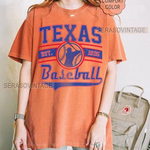 Pode incluir: Uma camiseta laranja queimada com um gr&aacute;fico de beisebol vintage. O gr&aacute;fico apresenta um jogador de beisebol em silhueta, uma bola de beisebol e o texto "Texas Baseball Est. 1835".