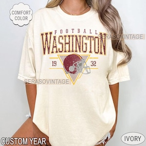 May include: Ivory-colored t-shirt with the word "WASHINGTON" in maroon and gold, above a football helmet graphic. The shirt also features the numbers "19" and "32". The words "COMFORT COLOR", "CUSTOM YEAR", and "IVORY" are also visible.