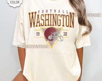 Op de afbeelding: Crèmekleurig T-shirt met het woord "WASHINGTON" in bordeauxrood en goud, boven een afbeelding van een footballhelm. Het shirt toont ook de nummers "19" en "32". De woorden "COMFORT COLOR", "CUSTOM YEAR" en "IVORY" zijn ook zichtbaar.