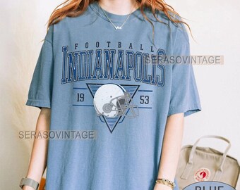 May include: A light blue t-shirt featuring the text "FOOTBALL INDIANAPOLIS" in navy blue. A white football helmet graphic is inside a navy blue triangle, with the numbers "19" and "53" on either side. The words "CUSTOM YEAR" and "BLUE JEAN" are also visible.