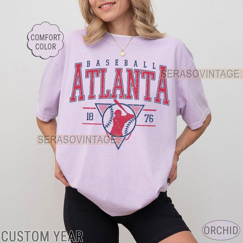 May include: A lavender t-shirt with a vintage baseball graphic. The graphic features a baseball player in a red and blue uniform with the text "BASEBALL ATLANTA 18 76".