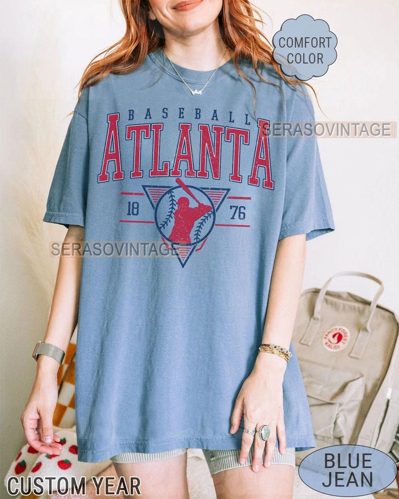 May include: A blue t-shirt with a vintage baseball design featuring the words "Baseball Atlanta" and the numbers "76" and "18" in red. The design also includes a baseball player in a red and white uniform. The shirt is a vintage style and has a distressed look.