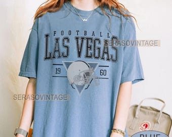 以下が含まれることがあります： 「FOOTBALL LAS VEGAS」の黒い文字がプリントされた水色のTシャツ。その下に、グレーのフットボールヘルメットが三角形の中に配置され、両側に数字の「19」と「60」があります。シャツには「CUSTOM YEAR」と「BLUE JEAN」の文字もプリントされています。
