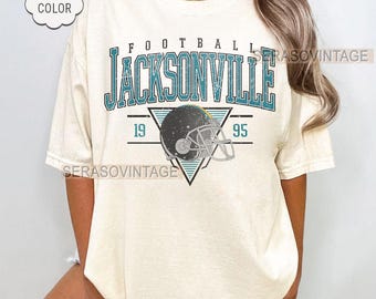 May include: Ivory t-shirt featuring a distressed teal and black graphic. The graphic reads "FOOTBALL JACKSONVILLE 1995" with a football helmet inside a triangle. The shirt has a comfort colour and custom year option.