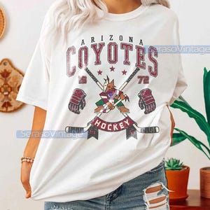Puede incluir: Camiseta blanca con un gr&aacute;fico de estilo vintage del equipo de hockey de los Arizona Coyotes. El gr&aacute;fico presenta el logotipo del equipo, palos de hockey cruzados y las palabras "Arizona Coyotes" y "Hockey".