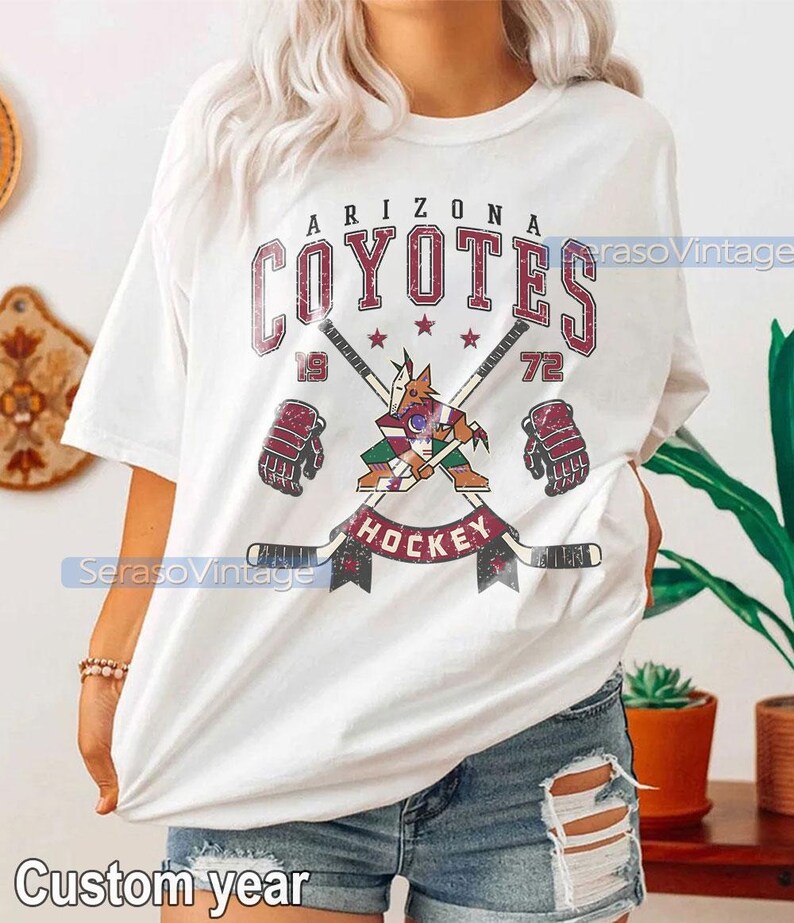 Puede incluir: Camiseta blanca con un gr&aacute;fico de estilo vintage del equipo de hockey de los Arizona Coyotes. El gr&aacute;fico presenta el logotipo del equipo, palos de hockey cruzados y las palabras "Arizona Coyotes" y "Hockey".