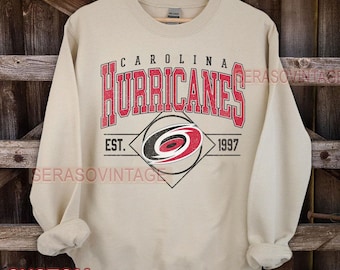 Puede incluir: Una sudadera de color beige con el logotipo de los Carolina Hurricanes y el texto "Carolina Hurricanes Est. 1997".