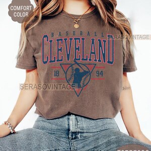 Puede incluir: Una camiseta marr&oacute;n con un gr&aacute;fico de b&eacute;isbol vintage que incluye el texto "BASEBALL CLEVELAND 18 94" y una silueta de un jugador de b&eacute;isbol. La camiseta la lleva una persona con el pelo largo y casta&ntilde;o y va combinada con unos vaqueros azules.