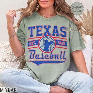 Pode incluir: Uma camiseta verde claro com um design gr&aacute;fico de estilo vintage que apresenta o texto "Texas Baseball" com uma silhueta de jogador de beisebol e o texto "Est. 1935".