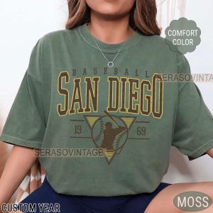 Może przedstawiać: Zielony T-shirt z vintage grafiką baseballową. Grafika przedstawia baseballistę z kijem i piłką oraz tekst "BASEBALL SAN DIEGO 19 69".