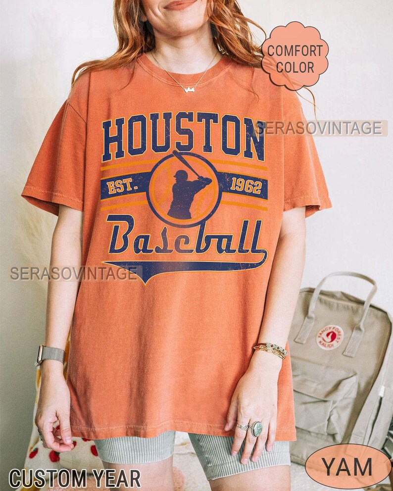 Puede incluir: Camiseta naranja con un dise&ntilde;o de b&eacute;isbol vintage. El dise&ntilde;o presenta el texto "HOUSTON Baseball EST. 1962" con una silueta de un jugador de b&eacute;isbol en el centro.