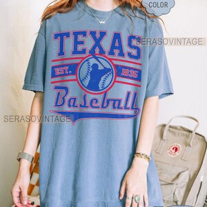 Pode incluir: Uma camiseta de jeans azul com o texto "Texas Baseball" em letras vermelhas e brancas. A camiseta tamb&eacute;m apresenta uma silhueta de jogador de beisebol e o texto "Est. 1835".
