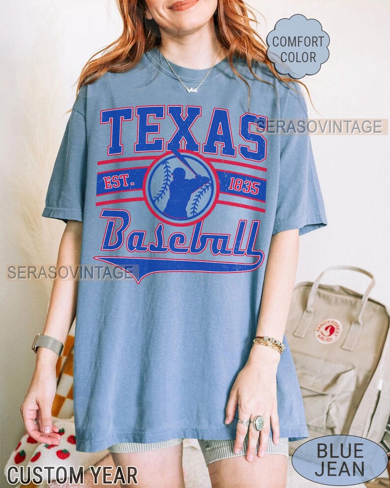 Pode incluir: Uma camiseta de jeans azul com o texto "Texas Baseball" em letras vermelhas e brancas. A camiseta tamb&eacute;m apresenta uma silhueta de jogador de beisebol e o texto "Est. 1835".