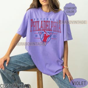 Puede incluir: Una camiseta morada con un gr&aacute;fico de b&eacute;isbol vintage. El gr&aacute;fico presenta a un jugador de b&eacute;isbol con un uniforme rojo y azul, una pelota de b&eacute;isbol y el texto "BASEBALL PHILADELPHIA 18 83".