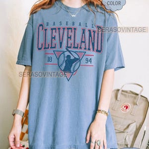 Puede incluir: Una camiseta azul con un gr&aacute;fico de b&eacute;isbol vintage que presenta la palabra "CLEVELAND" en letras rojas y blancas, una silueta de jugador de b&eacute;isbol y los n&uacute;meros "18" y "94". La camiseta tiene un aspecto desgastado y est&aacute; hecha de una tela suave y c&oacute;moda.