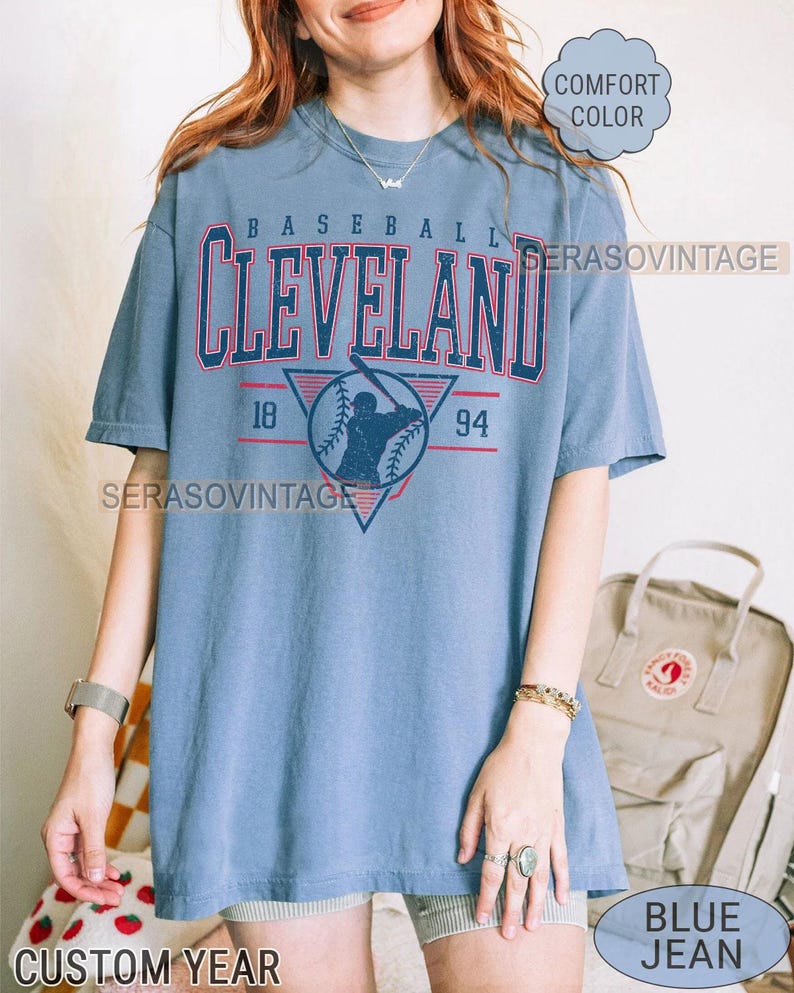 Puede incluir: Una camiseta azul con un gr&aacute;fico de b&eacute;isbol vintage que presenta la palabra "CLEVELAND" en letras rojas y blancas, una silueta de jugador de b&eacute;isbol y los n&uacute;meros "18" y "94". La camiseta tiene un aspecto desgastado y est&aacute; hecha de una tela suave y c&oacute;moda.