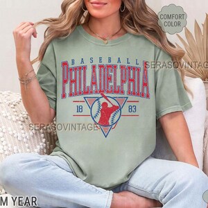 Puede incluir: Una camiseta verde salvia con un gr&aacute;fico de b&eacute;isbol vintage. El gr&aacute;fico presenta a un jugador de b&eacute;isbol en rojo y azul, con el texto "BASEBALL PHILADELPHIA 18 83".
