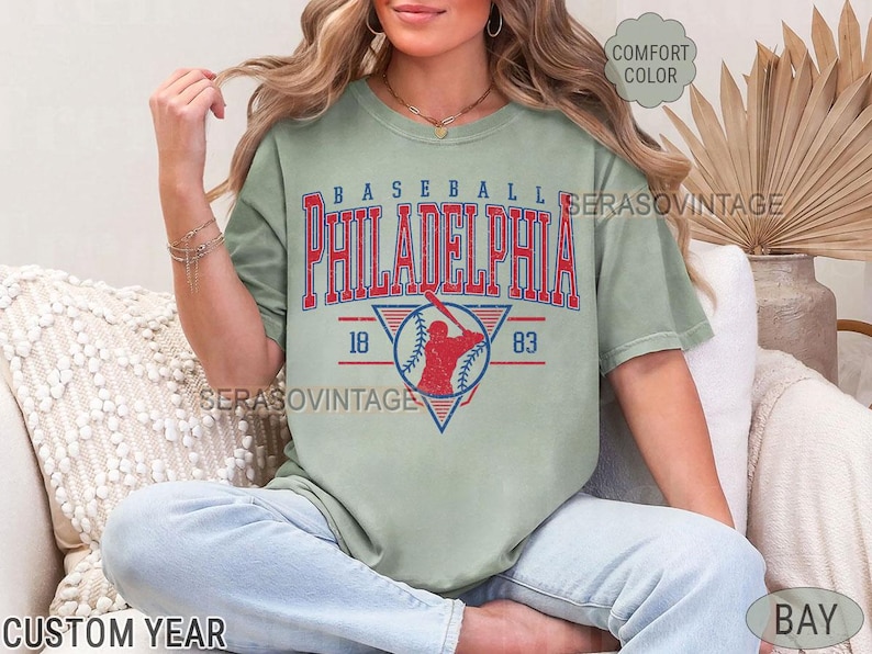Puede incluir: Una camiseta verde salvia con un gr&aacute;fico de b&eacute;isbol vintage. El gr&aacute;fico presenta a un jugador de b&eacute;isbol en rojo y azul, con el texto "BASEBALL PHILADELPHIA 18 83".