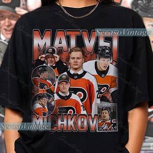 May include: Black t-shirt with a hockey-themed design. It features images of a hockey player, the name "MATVEI MICHKOV", and the number 39. The text is in orange and white.