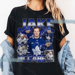 Könnte beinhalten: Schwarzes T-Shirt mit einer Grafik des Eishockeyspielers Jake McCabe im Trikot der Toronto Maple Leafs. Das Design enthält den Namen "JAKE" in Blau und Silber sowie mehrere Bilder des Spielers und das Teamlogo. Der Text "MCCABE" ist ebenfalls sichtbar.