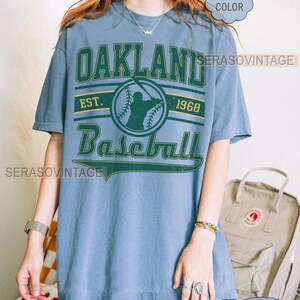 May include: A blue t-shirt with a vintage Oakland Baseball logo. The logo features a baseball player in silhouette, the words "Oakland Baseball", and the years "Est. 1968".