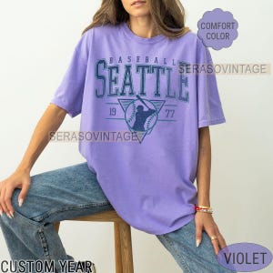 May include: A purple t-shirt with a vintage baseball graphic that says "Baseball Seattle 19 77".