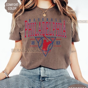Puede incluir: Una camiseta marr&oacute;n con un gr&aacute;fico de b&eacute;isbol vintage. El gr&aacute;fico presenta a un jugador de b&eacute;isbol en una silueta roja, una pelota de b&eacute;isbol y el texto "BASEBALL PHILADELPHIA 18 83".