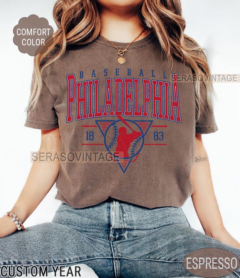 Puede incluir: Una camiseta marr&oacute;n con un gr&aacute;fico de b&eacute;isbol vintage. El gr&aacute;fico presenta a un jugador de b&eacute;isbol en una silueta roja, una pelota de b&eacute;isbol y el texto "BASEBALL PHILADELPHIA 18 83".