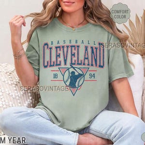 Puede incluir: Una camiseta verde con un gr&aacute;fico de b&eacute;isbol vintage. El gr&aacute;fico presenta a un jugador de b&eacute;isbol en un tri&aacute;ngulo con el texto "BASEBALL CLEVELAND 18 94".