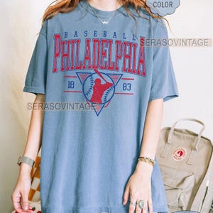 Puede incluir: Una camiseta vintage azul con el texto "BASEBALL PHILADELPHIA" en letras rojas y blancas. La camiseta presenta un gr&aacute;fico de jugador de b&eacute;isbol con los a&ntilde;os "18" y "83" a cada lado del gr&aacute;fico. La camiseta la lleva puesta una persona con pantalones cortos de mezclilla azul y un reloj plateado.