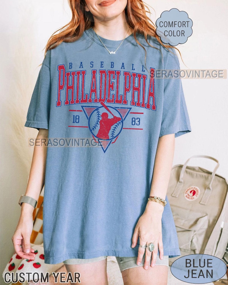 Puede incluir: Una camiseta vintage azul con el texto "BASEBALL PHILADELPHIA" en letras rojas y blancas. La camiseta presenta un gr&aacute;fico de jugador de b&eacute;isbol con los a&ntilde;os "18" y "83" a cada lado del gr&aacute;fico. La camiseta la lleva puesta una persona con pantalones cortos de mezclilla azul y un reloj plateado.