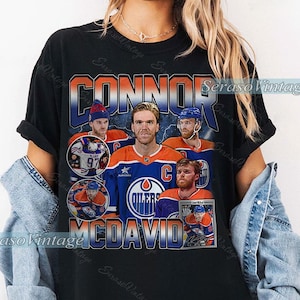May include: Black t-shirt featuring a vintage-style graphic of Connor McDavid, an ice hockey player. The design includes images of McDavid in his Oilers uniform, with the name "CONNOR MCDAVID" in bold lettering. The shirt has a retro aesthetic.