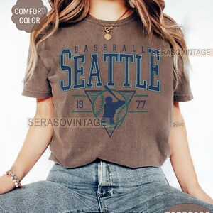 May include: A brown t-shirt with a vintage baseball graphic featuring the text "Seattle" and the numbers "19" and "77".