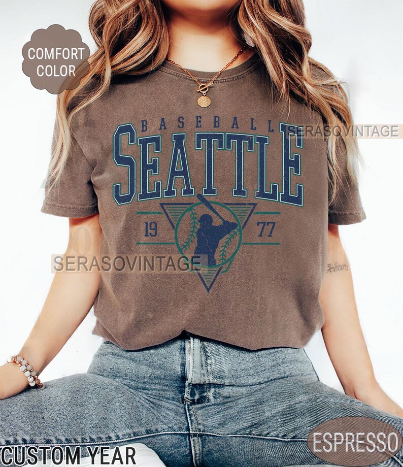 May include: A brown t-shirt with a vintage baseball graphic featuring the text "Seattle" and the numbers "19" and "77".