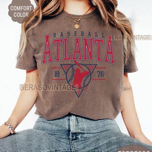 May include: A brown t-shirt with a vintage baseball graphic. The graphic features a baseball player in a red silhouette, a baseball, and the text "BASEBALL ATLANTA 76 18".