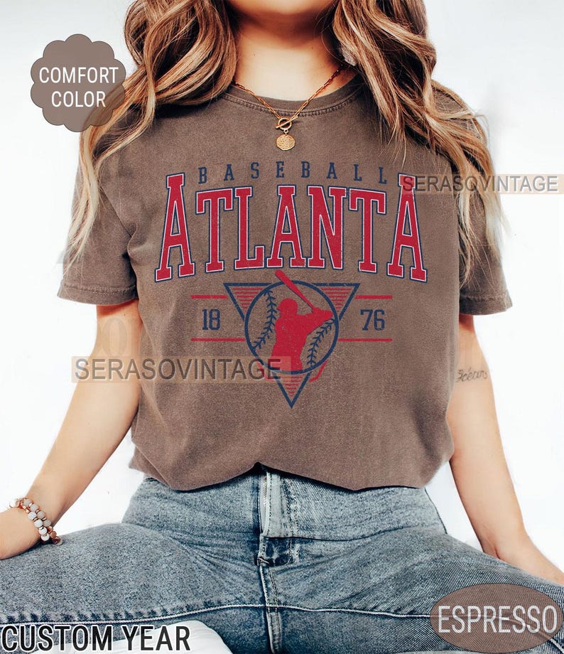 May include: A brown t-shirt with a vintage baseball graphic. The graphic features a baseball player in a red silhouette, a baseball, and the text "BASEBALL ATLANTA 76 18".