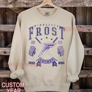 Peut inclure: Un sweat-shirt beige avec un motif graphique violet et blanc. Le graphique présente le texte "Minnesota Frost Hockey" avec des bâtons de hockey croisés et un éclair. Les années 20 et 23 sont également incluses dans le design.