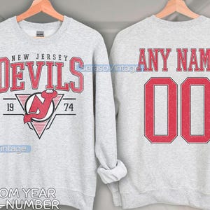 May include: Two gray sweatshirts, one with the New Jersey Devils logo and the text "NEW JERSEY DEVILS 19 74" and the other with the text "ANY NAME 00".