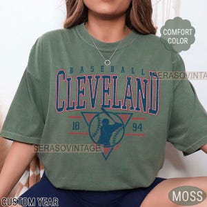 Puede incluir: Una camiseta verde con un gr&aacute;fico de b&eacute;isbol vintage. El gr&aacute;fico presenta a un jugador de b&eacute;isbol en silueta, una pelota de b&eacute;isbol y el texto "BASEBALL CLEVELAND 18 94".